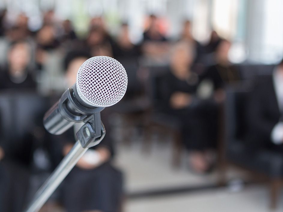 Ein Mikrofon im Vordergrund und im Hintergrund sitzen Menschen in schwarzer Kleidung.